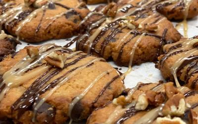 Dozens of cookies cooling off on parchment paper, topped with pecans and drizzled with caramel and chocolate.