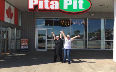 Two Pita Pit employees stand in front of the storefront with their arms up in the air, the Pita Pit sign above them.